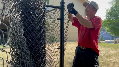 A worker in a red shirt and gloves secures a chain-link property fence to a metal post in a sunny outdoor area with buildings in the background.
