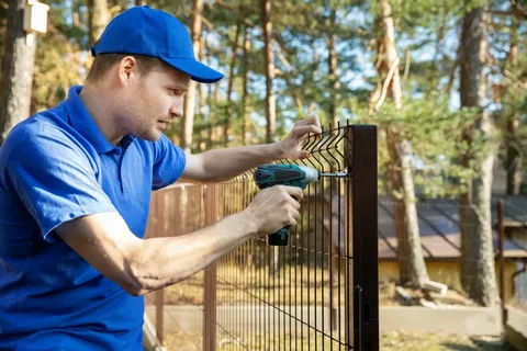 A maintenance worker in a blue uniform uses a power drill to secure a black metal property fence beside a wooded yard on a sunny day.